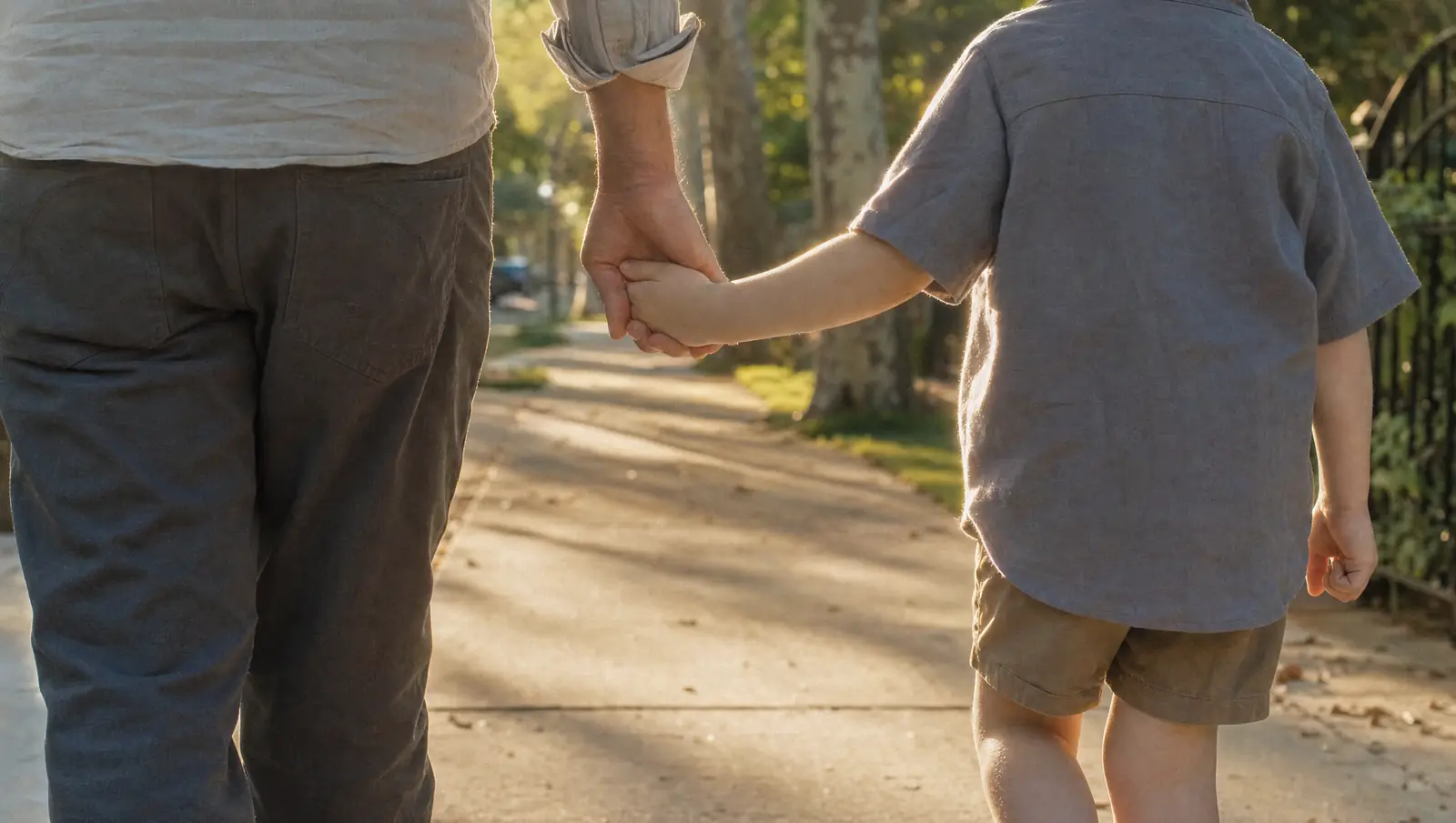 Seen from behind, a U visa applicant holding a small child's hand as they walk along a sunlit, tree-lined residential sidewalk.