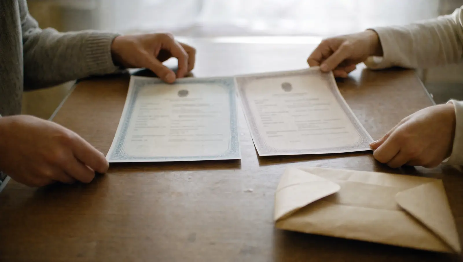 Two adults' hands laying their birth certificates for a sibling petition side by side on a desk next to a receipt envelope