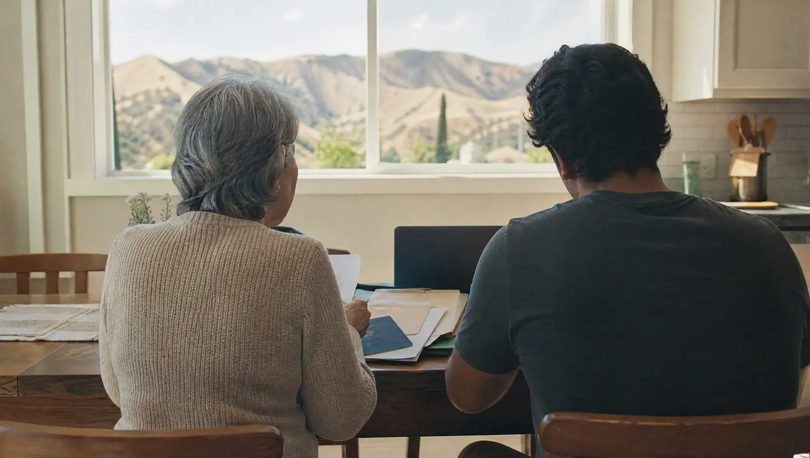 A Simi Valley family seen from behind sorting documents and a passport beside a laptop video call with their immigration lawyer, dry hills through the window