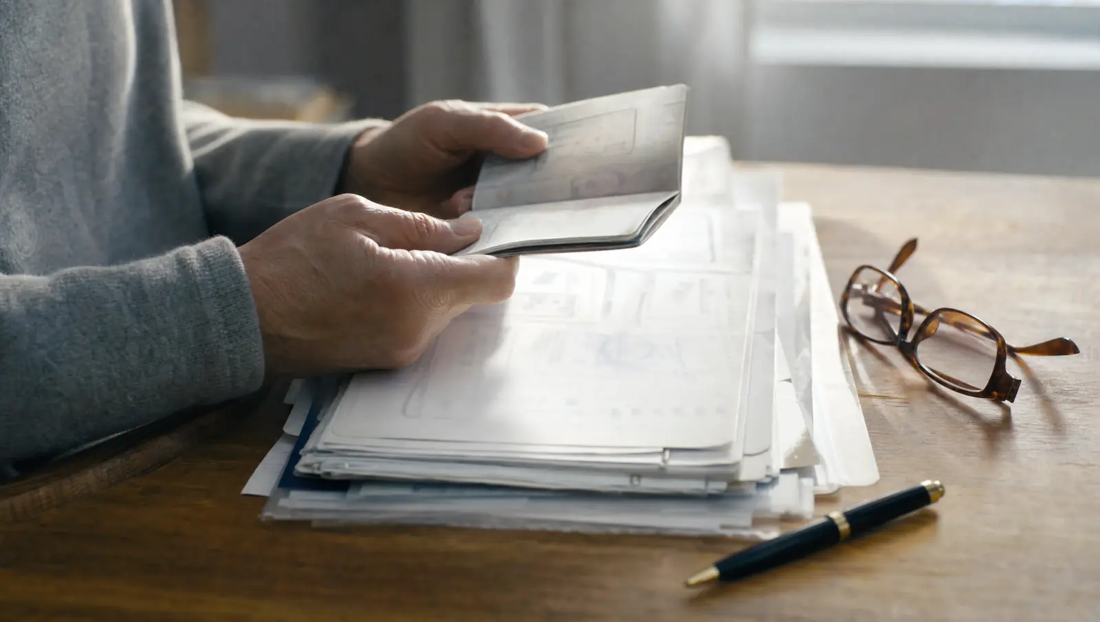 Two hands holding a passport open above a tidy stack of medical records and signed letters for an immigration waiver, with a pen and reading glasses beside them.