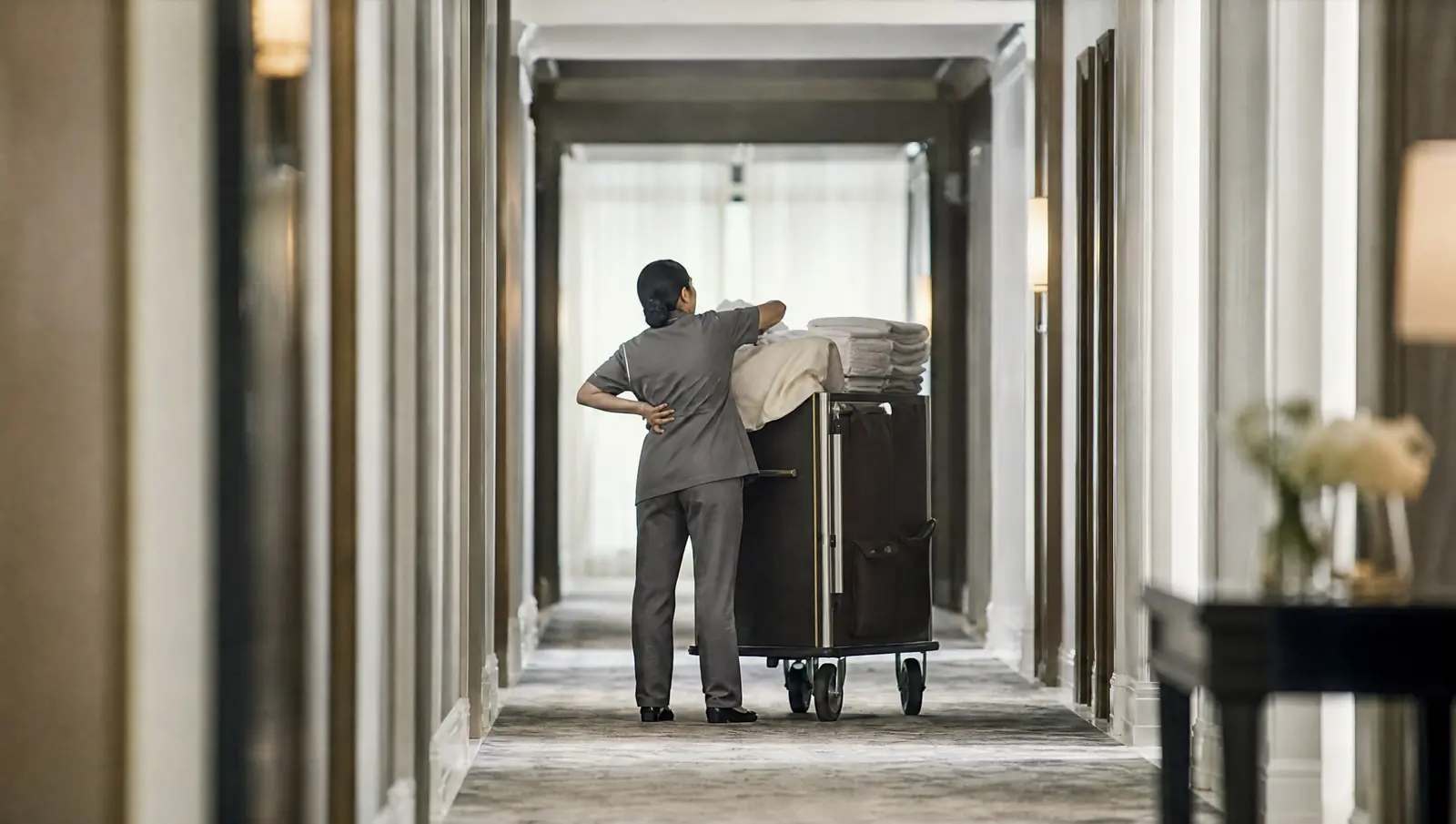 A Los Angeles hotel housekeeper seen from behind lifting a heavy bundle of linens beside a housekeeping cart, one hand pressed to her lower back, a common workers' comp injury
