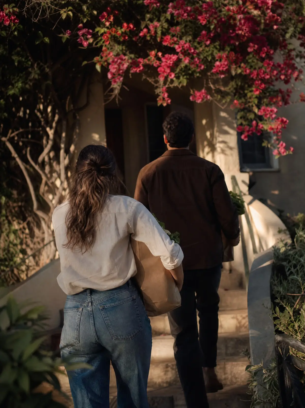 Two adults seen from behind carrying grocery bags up the steps of a small apartment building in afternoon sun