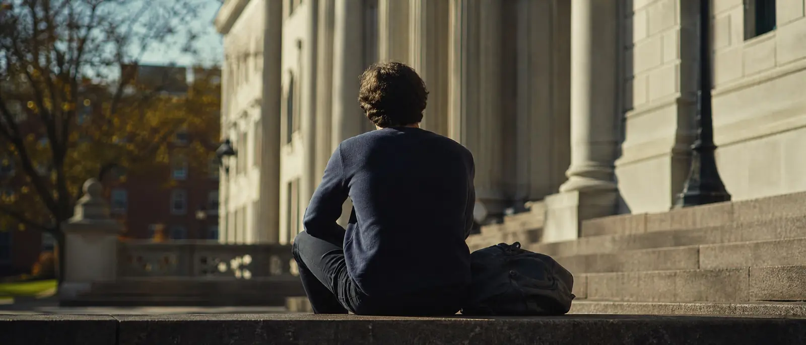 A young adult seen from behind sitting on university library steps in afternoon sun