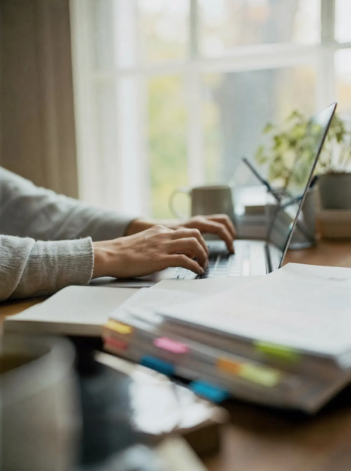 Hands typing on a laptop beside printed brief pages marked with coloured sticky flags at a bright desk