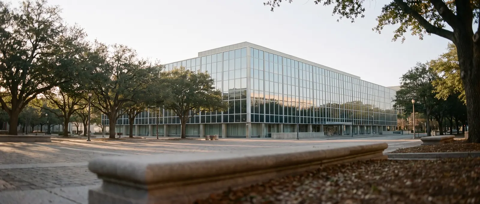 A modern office building with a glass facade reflecting blue sky, seen across a tree-lined plaza