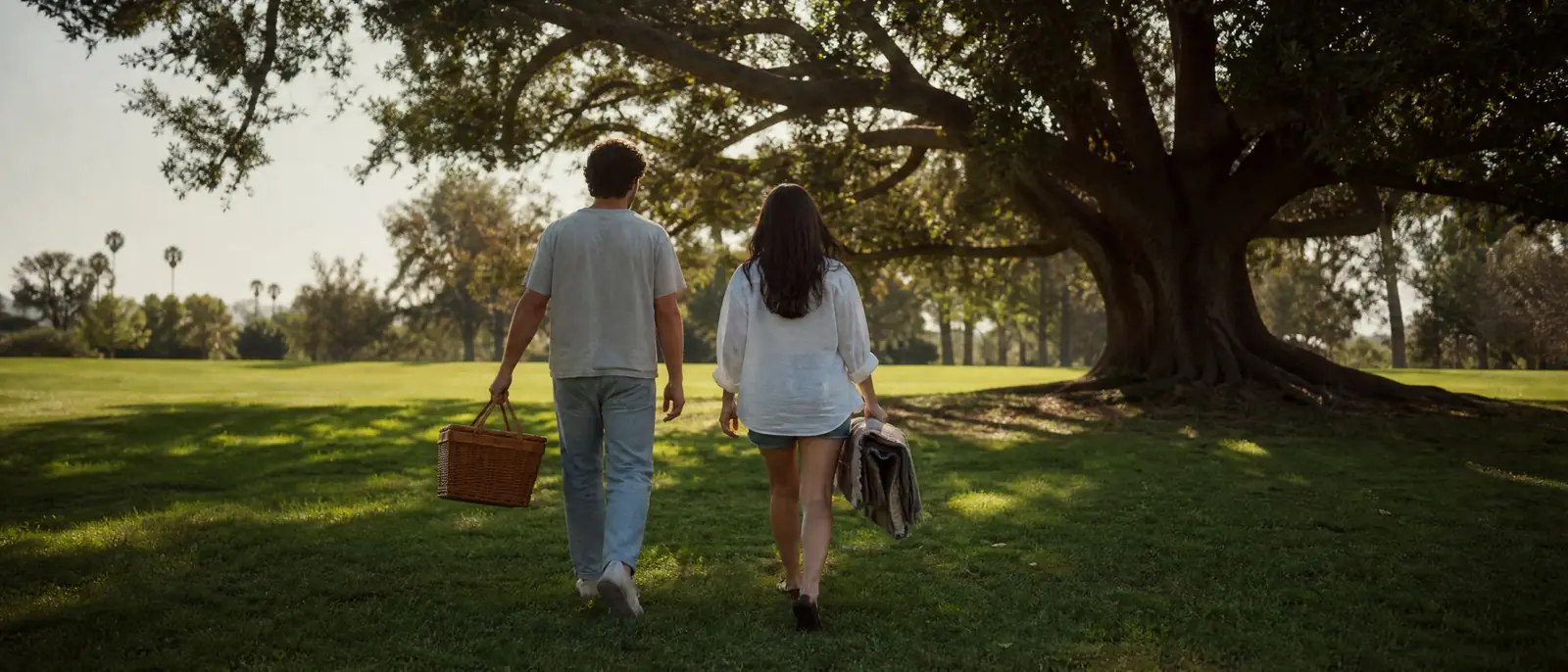 A young couple seen from behind carrying a picnic basket and blanket across a sunny park lawn toward a large shade tree