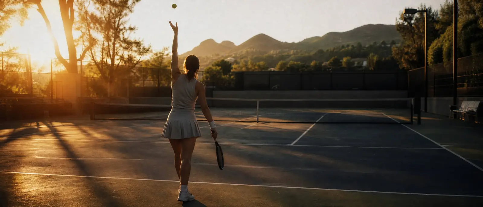 A tennis player seen from behind tossing the ball to serve on an empty outdoor court at golden hour