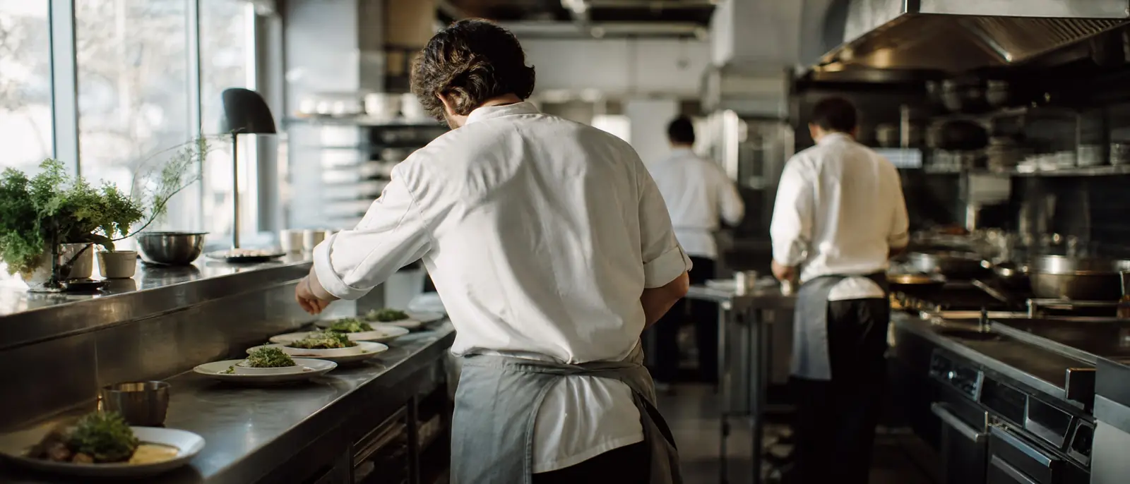 A chef seen from behind plating dishes along a steel pass in a bright restaurant kitchen during lunch service