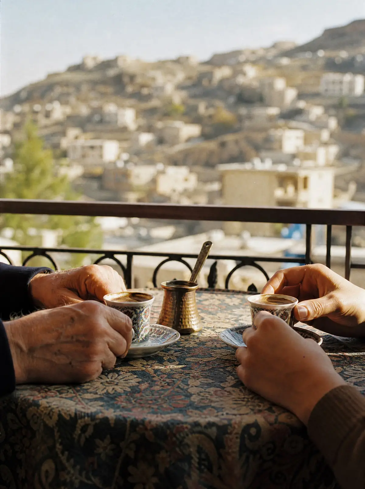 Two pairs of hands, one older and one younger, sharing small cups of Armenian coffee at a balcony table, hillside houses out of focus beyond