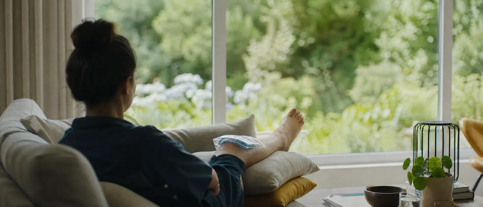 A person seen from behind resting on a sofa with an ice pack on a raised knee, looking out at a sunlit garden.