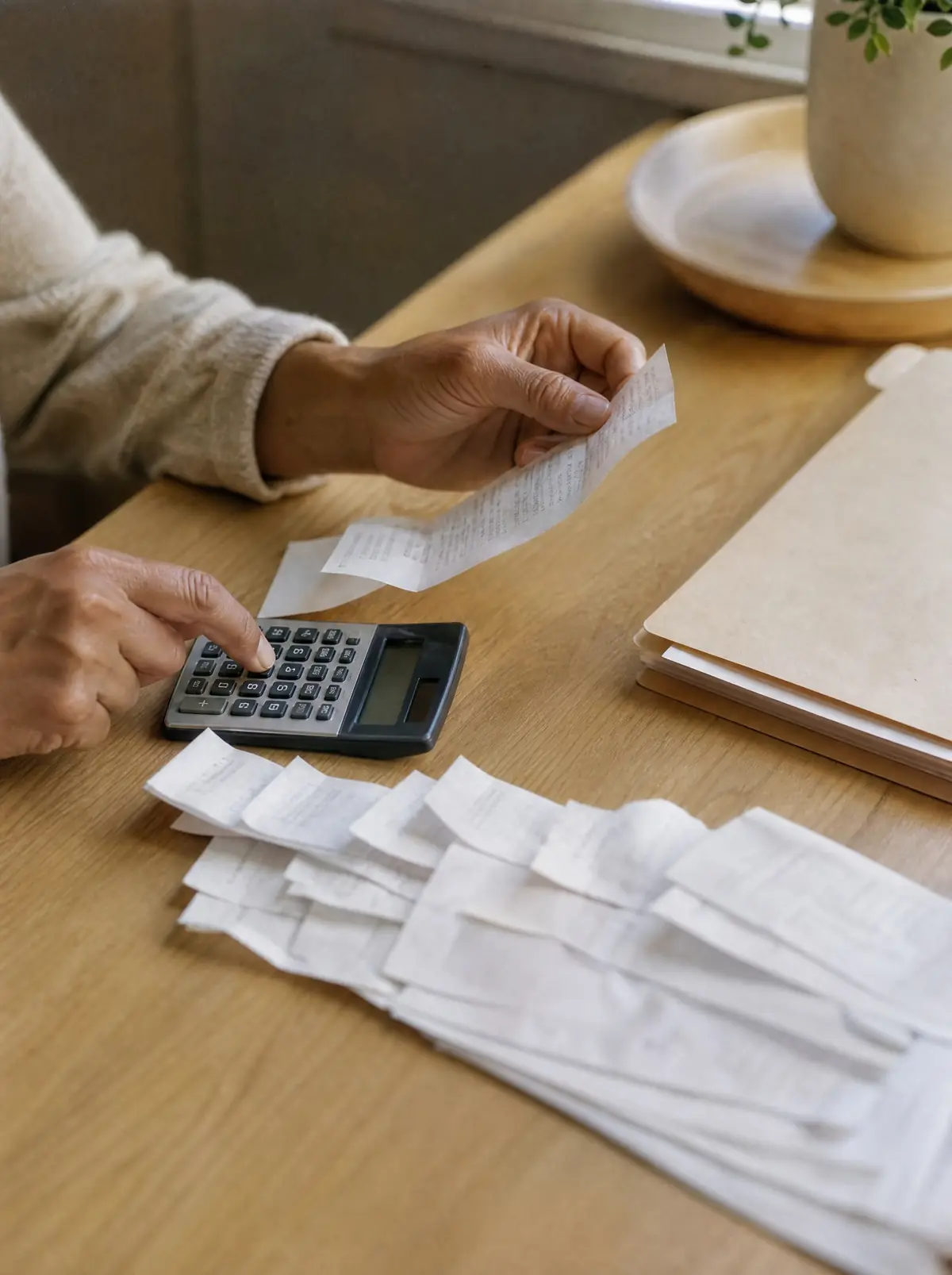 Two hands tallying medical receipts with a calculator beside a manila folder on a light oak desk.