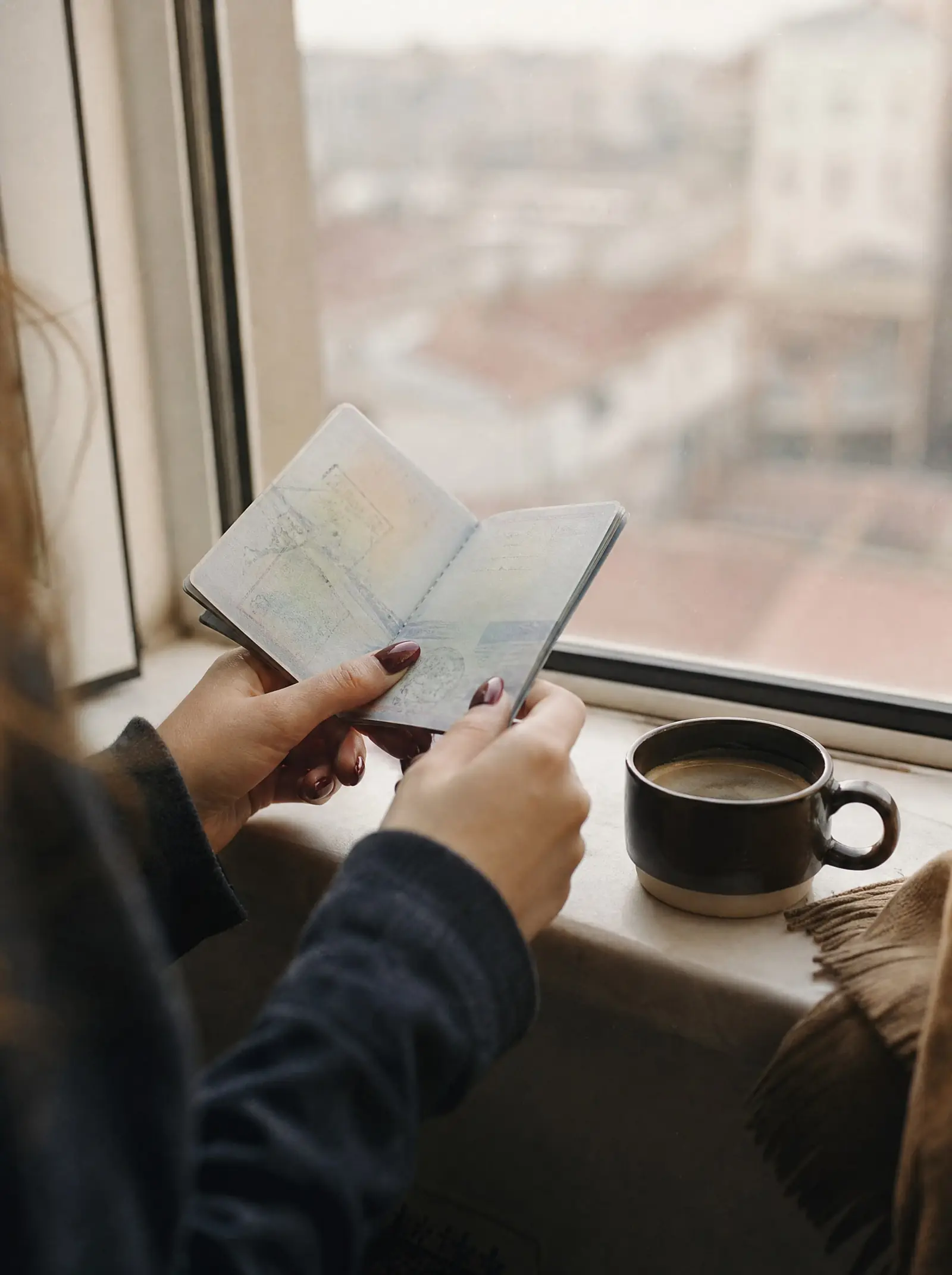 A woman's hands holding an old passport open to a visa page beside a cup of coffee on a sunlit windowsill